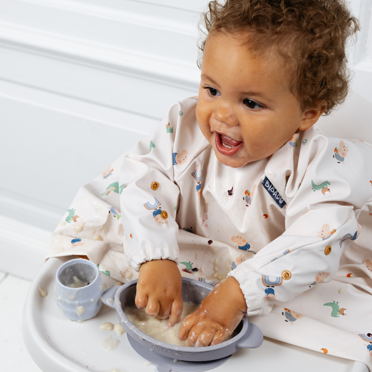 Happy baby in a high chair, wearing a patterned bib, playing with food in a light blue silicone suction bowl. - Product image