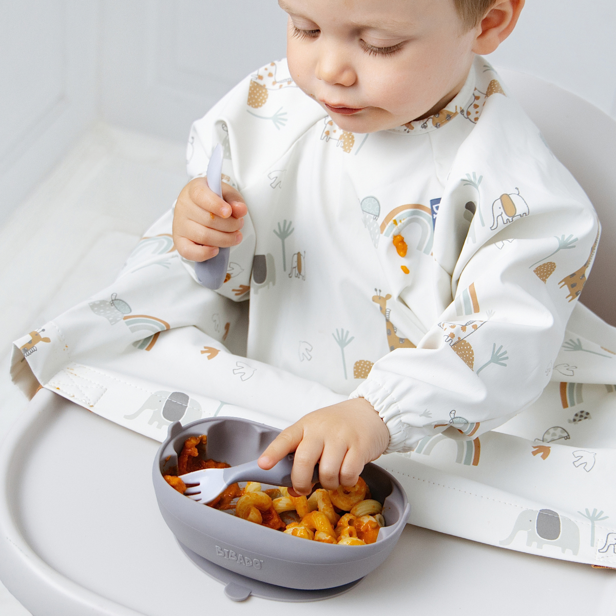 A baby wearing a white long-sleeve bib with a safari animal print sits in a high chair, using a blue silicone suction bowl and matching spoon to eat pasta. - Product info image
