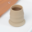 A beige silicone open cup with a matching base ring, shown on a white surface with part of an orange-brown sleeve visible in the background.