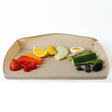 A beige silicone baby feeding mat with optional sides pulled up, shown on a clean white background and filled with colourful fruits, vegetables, and a small egg half.
