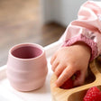 Pink silicone Sippit training cup next to a baby's hand reaching for a strawberry on a bamboo plate.