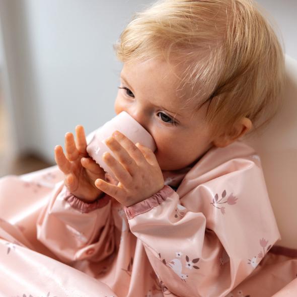 Baby drinking from a small, blush pink silicone open training cup, wearing a matching floral bib. - Product image