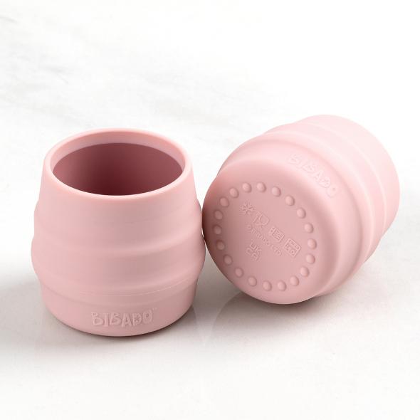 Two small, light pink silicone Sippit™ open training cups, one upright, one on its side revealing a textured base. - Product image