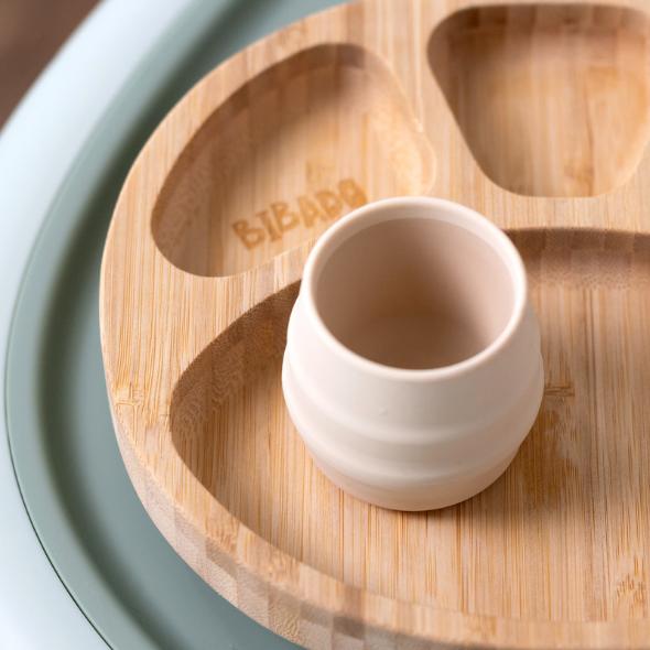 Bibado Sippit™ silicone open training cup in a light beige color, ribbed, on a wooden divided plate. - Product image