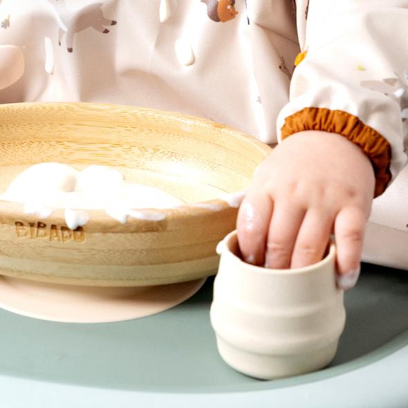 Baby's hand in a small, ribbed, beige silicone Sippit™ open training cup next to a wooden bowl. - Product image