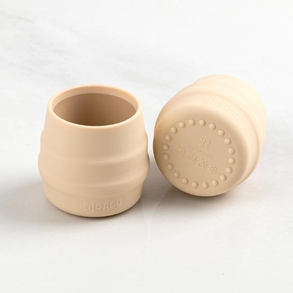 Two small, beige silicone Sippit™ open training cups with ribbed sides and a textured base. - Product image