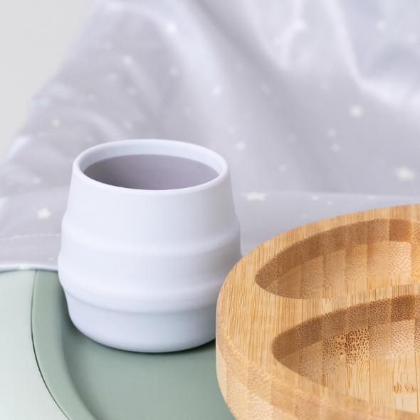 White silicone Sippit™ open training cup with ribbed design next to a wooden plate. - Product image