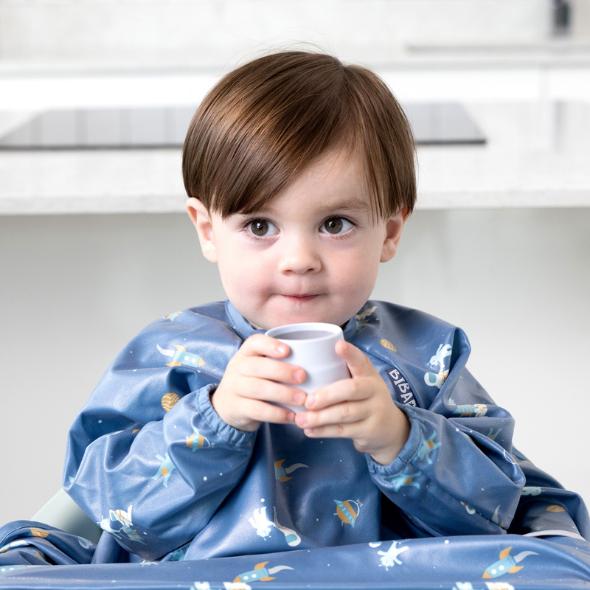Toddler in a blue space-themed bib holding a small, light gray silicone Sippit training cup. - Product image