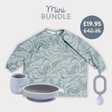 Pale green long-sleeved dinosaur print bib, grey silicone suction bowl, matching cup, and teething toy.