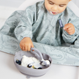 A baby wearing a light blue dinosaur-print bib sits in a high chair, reaching into a matching silicone bowl with a spoon while eating yogurt and blueberries.