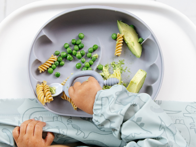 A baby wearing a light blue dinosaur-print bib sits in a high chair, reaching for food on a blue divided plate. The plate contains pasta, green peas, and slices of avocado. - image