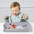 A toddler wearing a light blue dinosaur-print bib sits at a table with a blue-grey feeding placemat in front of them, eating strawberries and yoghurt with a spoon.