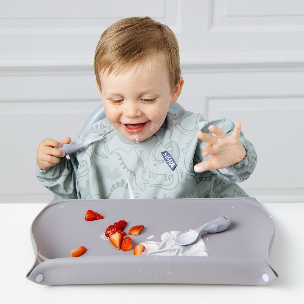 A toddler wearing a light blue dinosaur-print bib sits at a table with a blue-grey feeding placemat in front of them, eating strawberries and yoghurt with a spoon. - Product image