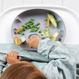 A baby wearing a light blue dinosaur-print bib sits in a high chair, reaching for food on a blue scalloped silicone plate. The plate contains pasta, green peas, and slices of avocado.