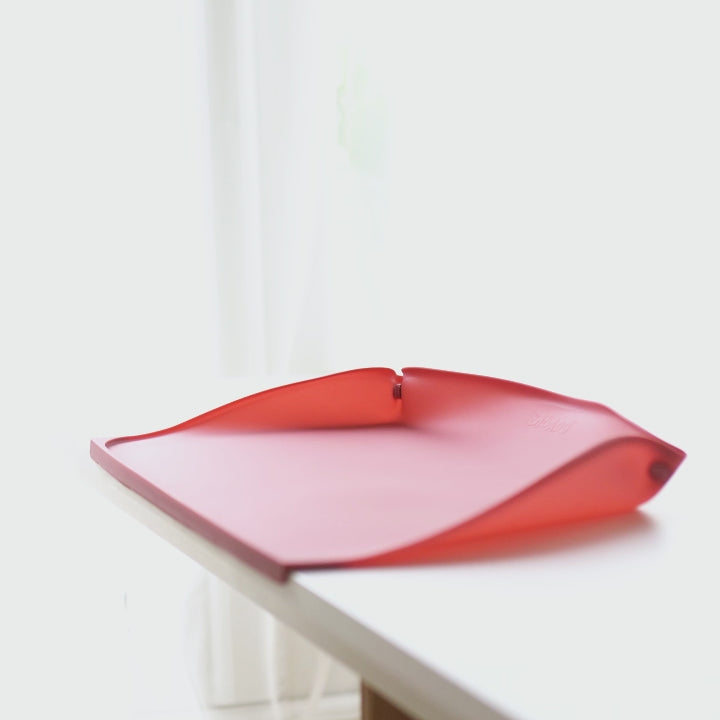 A toddler wearing a light blue dinosaur-print bib sits at a table with a blue-grey feeding placemat in front of them, eating strawberries and yoghurt with a spoon. - Product image