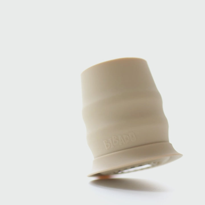 A gray silicone baby feeding cup with measurement markings inside, shown on a white surface with a patterned bib in the background. - Product image
