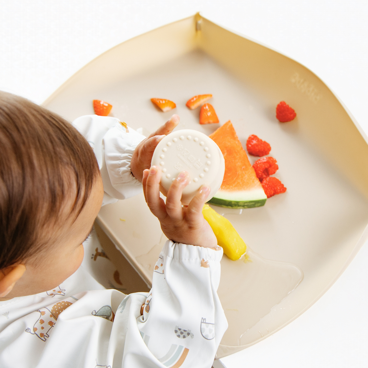 A baby sits in front of a beige feeding mat with the sides raised, spilling a small cup full of water onto the mat. The mat is styled with fruit pieces (watermelon, mango, strawberries) and a small beige cup, set against a clean white background. - Product image