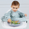 A baby wearing a light blue dinosaur-print bib sits in a white high chair, reaching for food on a matching blue plate.