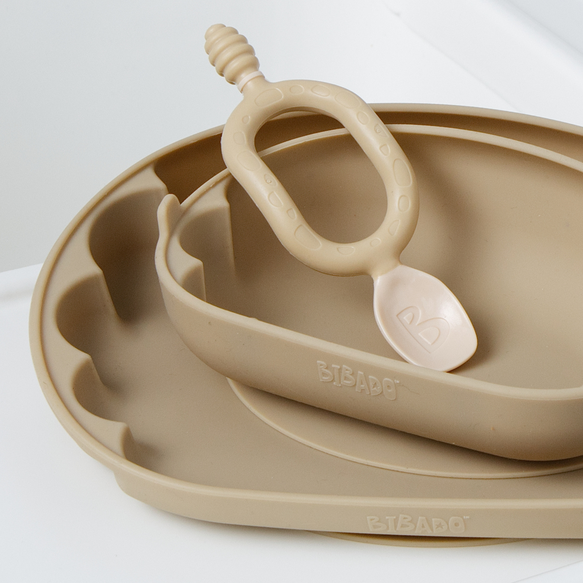 A beige silicone baby feeding set including a scalloped plate, a smaller bowl, and a Dippit spoon shown on a clean white background. The brand name 'Bibado' is embossed on the side of the bowl. - product info image
