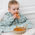Baby in a light green dinosaur-patterned long-sleeve bib, eating from a beige silicone suction bowl with a matching spoon.