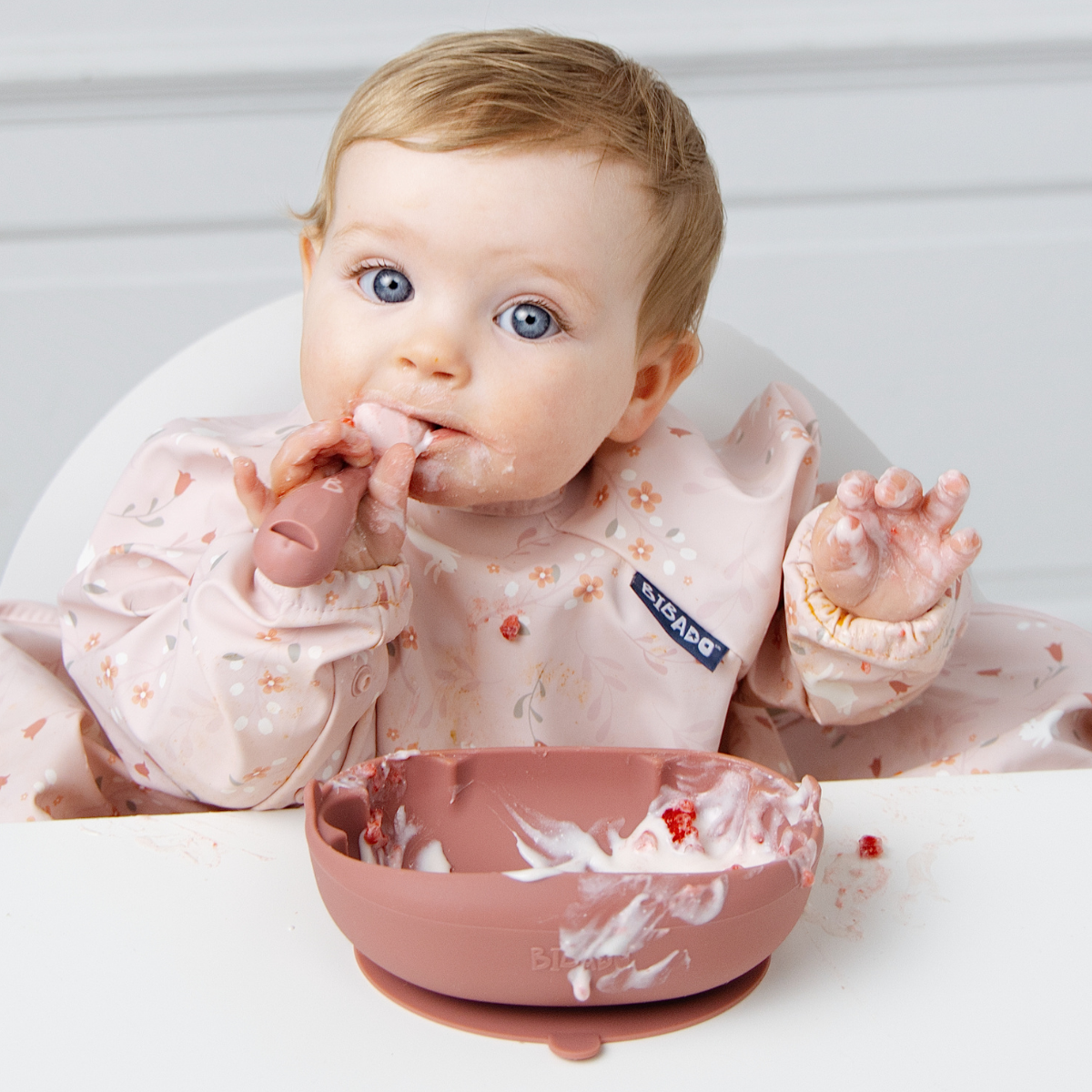 A baby wearing a light pink floral bib sits in a white high chair, eating from a dusty pink silicone bowl.  - Product image
