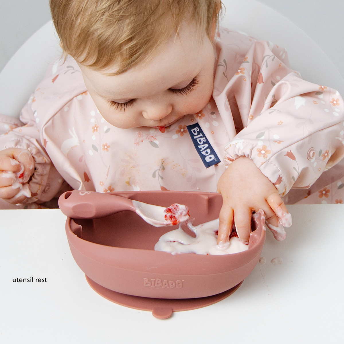 A baby wearing a light pink floral bib leans over a dusty rose silicone feeding bowl with a suction base, reaching for yoghurt in the bowl.  - Product image