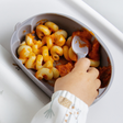 A child's hand wearing a white long-sleeve bib with animal print is using a blue Dippit spoon to eat macaroni pasta from a blue silicone feeding bowl. The bowl is filled with orange-colored pasta sauce and is set on a white high chair.