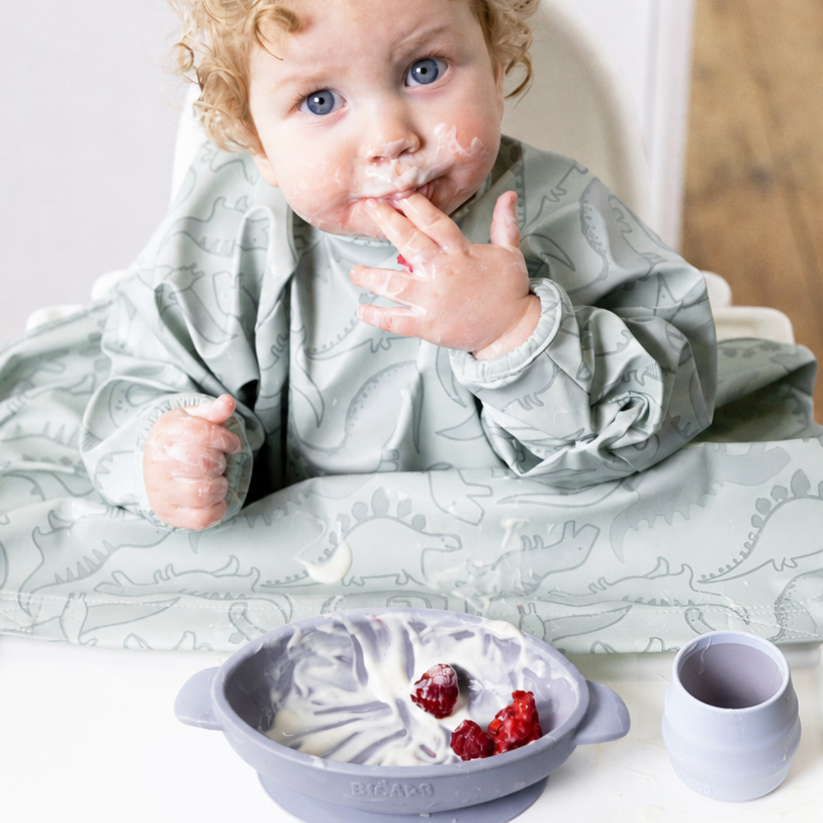 Messy baby with curly hair in a dinosaur bib, looking up, next to a mist silicone suction bowl with yogurt and berries. - Product image