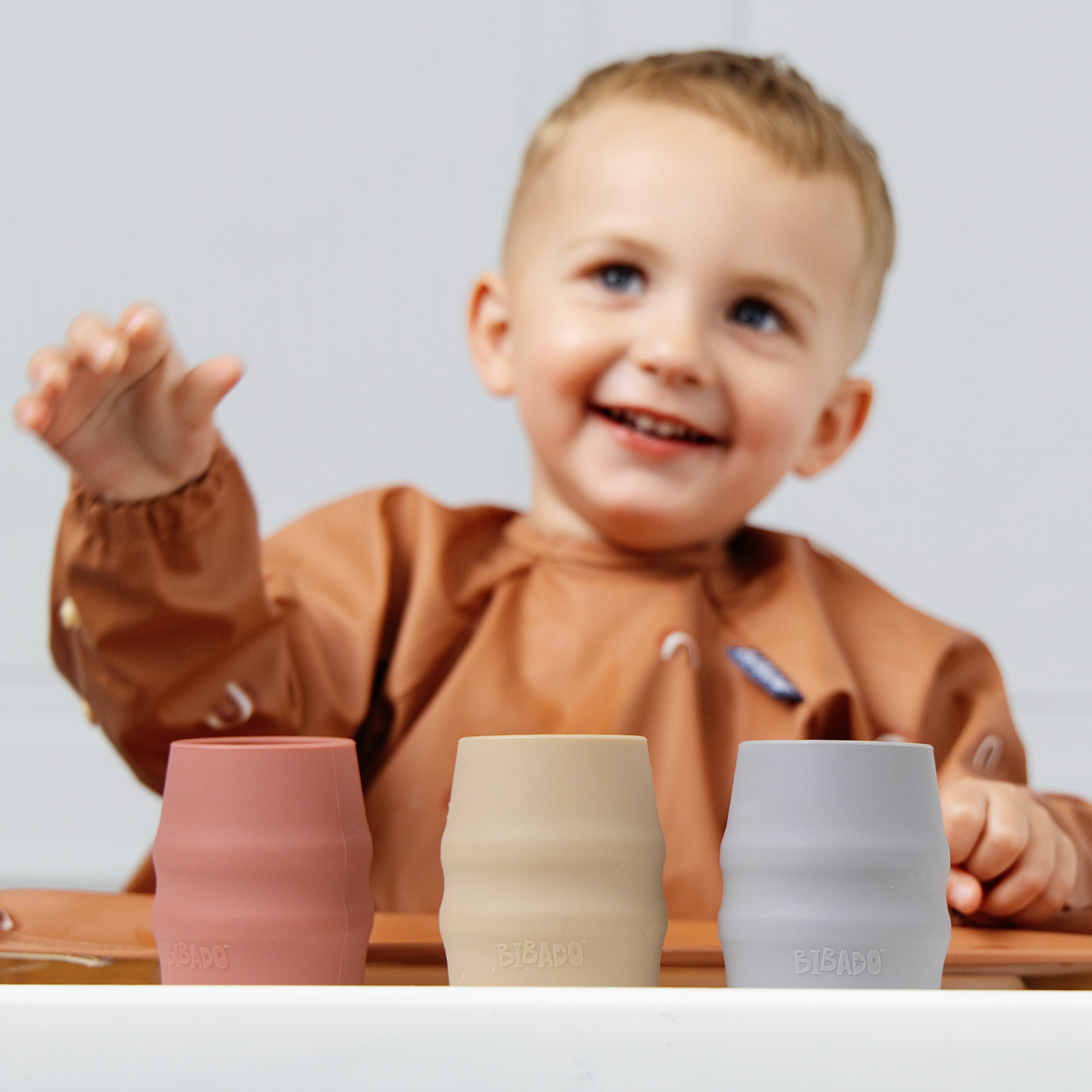 A smiling toddler in a brown bib reaches toward three silicone drinking cups on a tray. The cups are in dusty pink, warm beige, and light blue-grey. - Product image