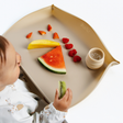 A baby sits in front of a beige feeding mat with the sides raised holding a kiwi slice. The mat is styled with fruit pieces (watermelon, mango, strawberries) and a small beige cup, set against a clean white background.