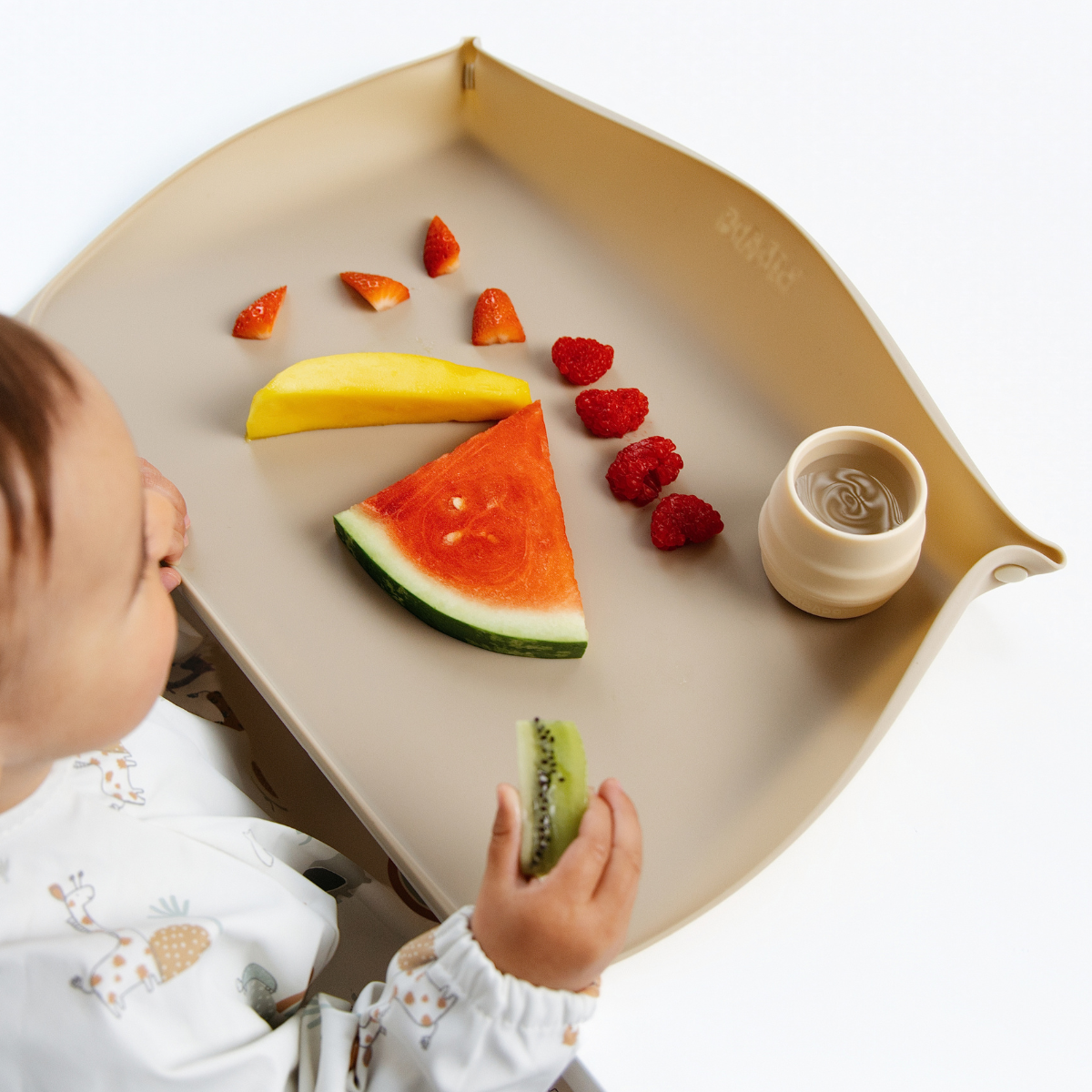 A baby sits in front of a beige feeding mat with the sides raised holding a kiwi slice. The mat is styled with fruit pieces (watermelon, mango, strawberries) and a small beige cup, set against a clean white background. - Product image
