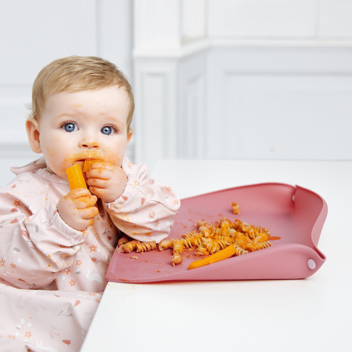 A baby wearing a light pink floral Coverall bib sits in a white high chair, eating a carrot with pasta on a pink feeding mat with the optional sides raised. The baby's face and hands are covered in orange food residue. - Product image