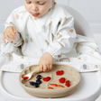 A baby wearing a white long-sleeve bib with a safari animal and rainbow print sits in a high chair, using a beige silicone plate to eat strawberries and blueberries.