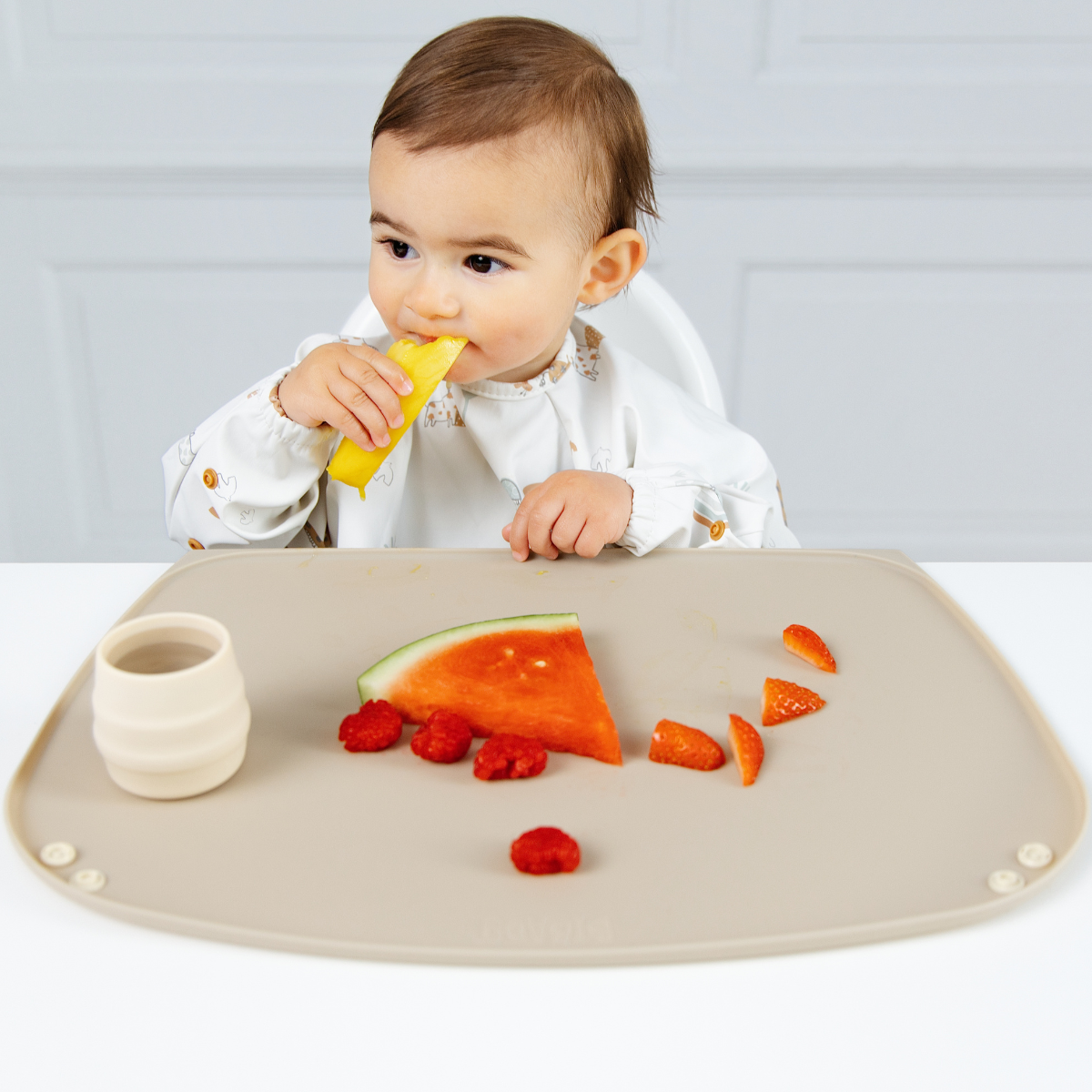 A baby wearing a white Coverall bib sits at a table using a beige silicone baby feeding placemat. On the mat rests a slice of watermelon, strawberries, and a small cream-colored Sippit cup. - Product image