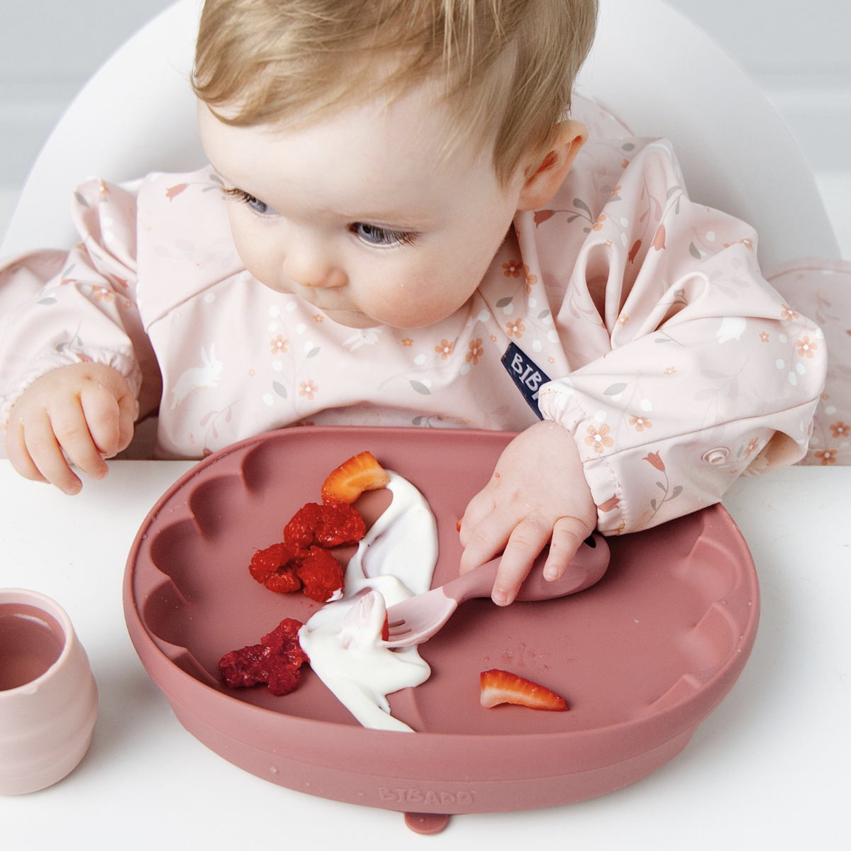A baby wearing a light pink floral bib sits in a white high chair, looking down at a dusty rose silicone baby plate with a spoon and food pieces. A small matching cup is visible on the left. - Product image