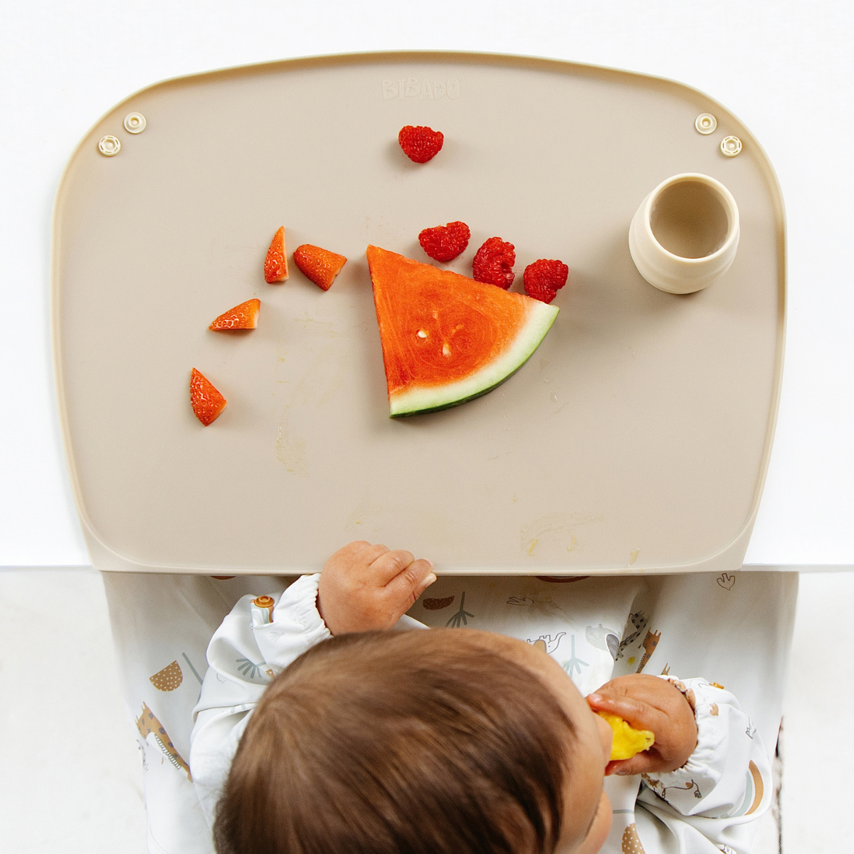 A baby sits at a white table with a beige silicone feeding mat, eating a slice of watermelon and mango. The tray has a small cream-colored cup sat on the right side. - Product image